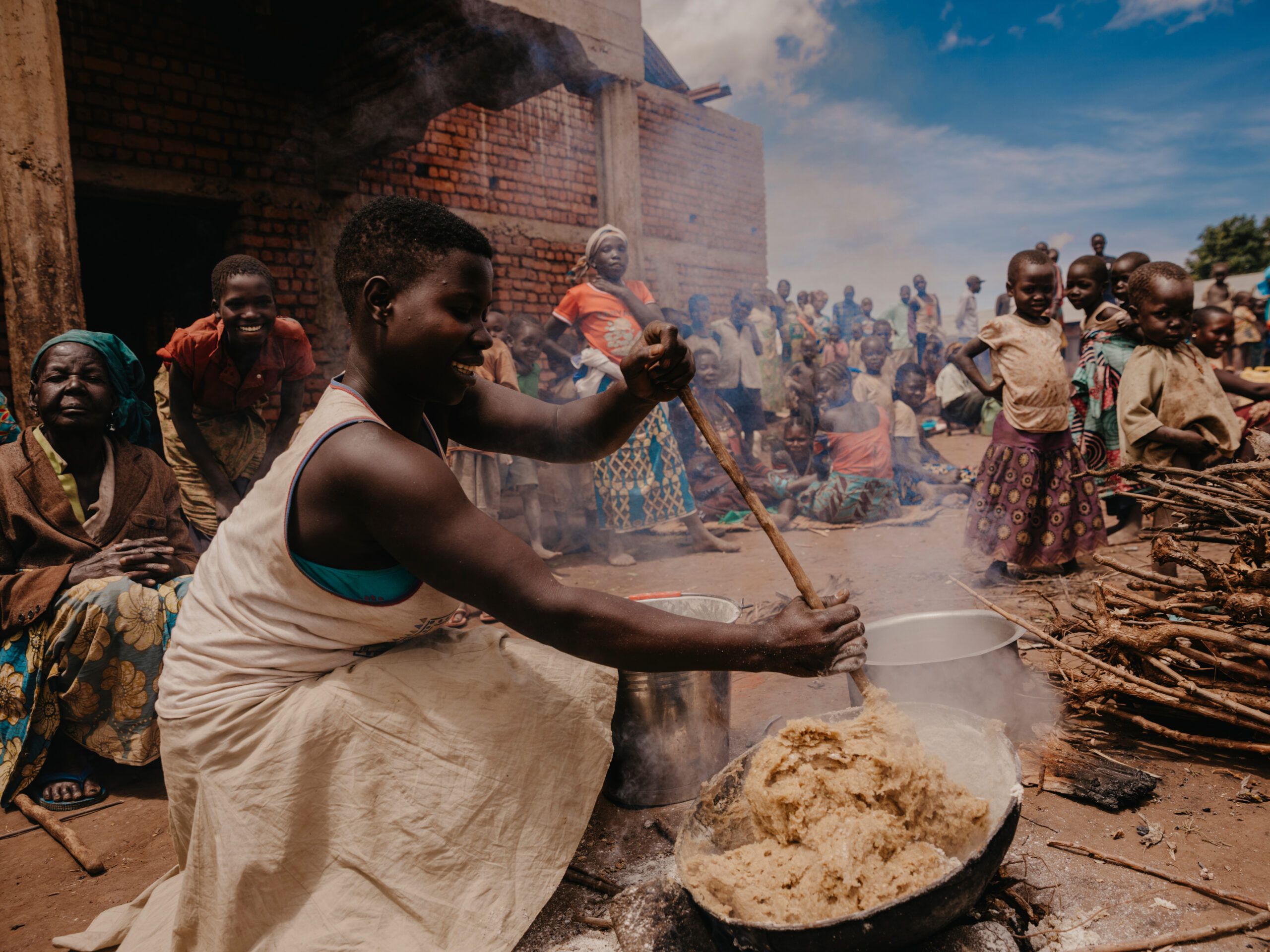 En afrikansk kvinne rører i en deig i en bålpanne. Rundt henne står flere barn og kvinner. I bakgrunnen skimtes også noen menn.