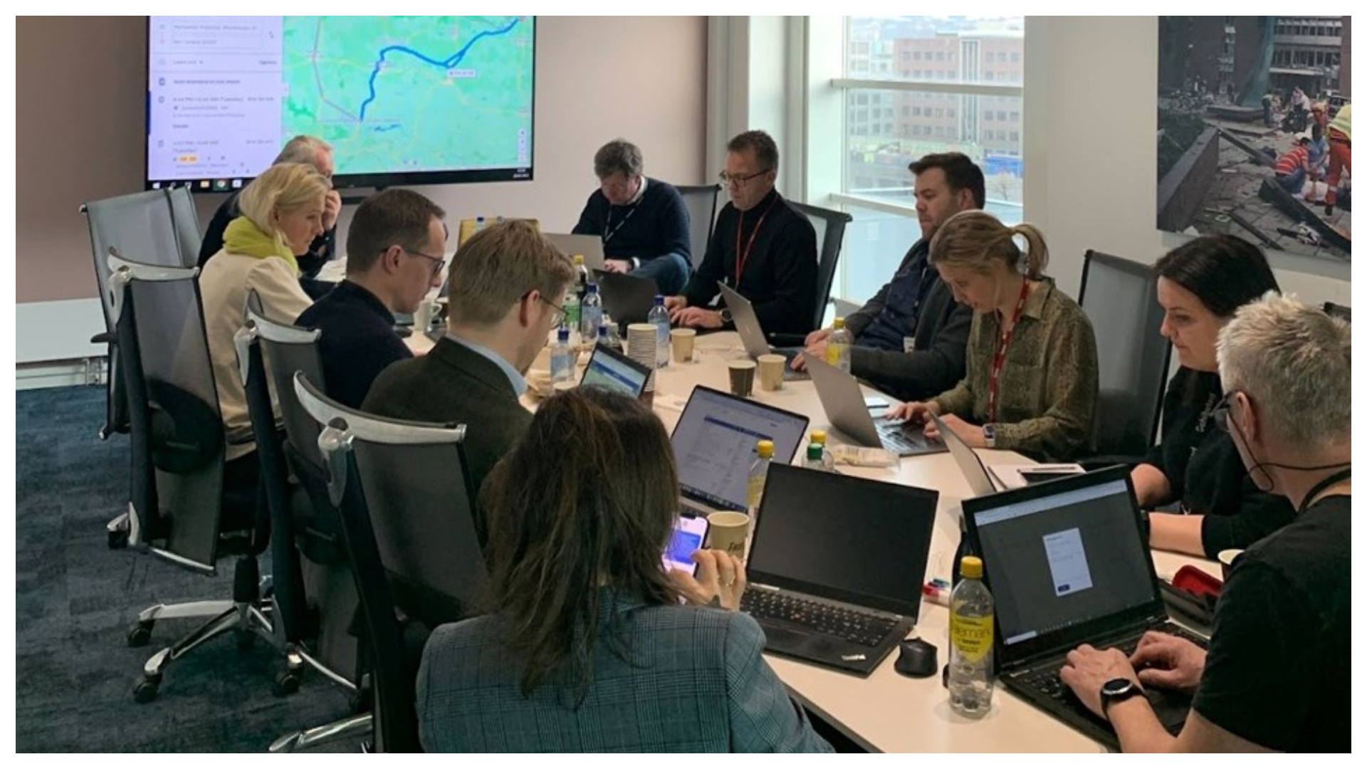 Mennesker som sitter rundt et bord i et møterom. De fleste ser ned i pc-skjermene sine. På veggen er det en skjerm hvor vi skimter et kart.