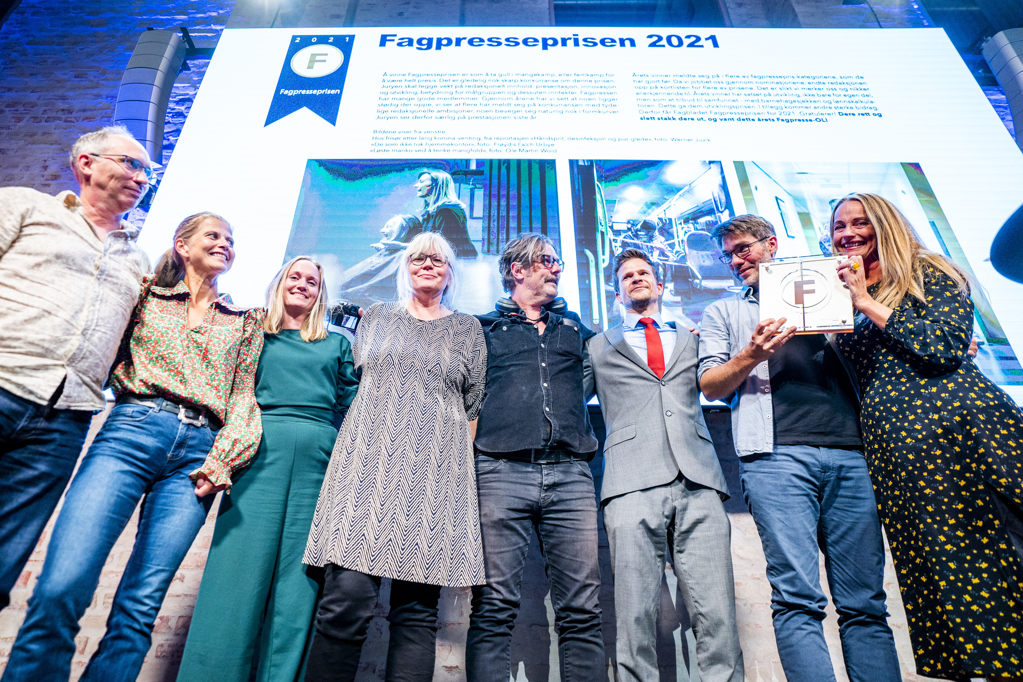 Glade prisvinnere står på rekke og holder rundt hverandre, foran en storskjerm med påskriften "Fagpresseprisen 2021"